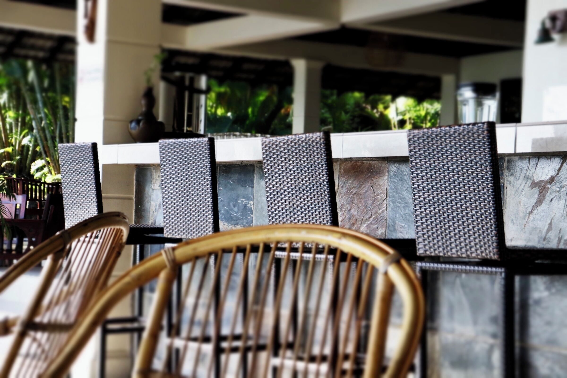 A seating area with a wicker chair and a metal chair, a stone wall with a textured pattern, and a view of greenery outside.