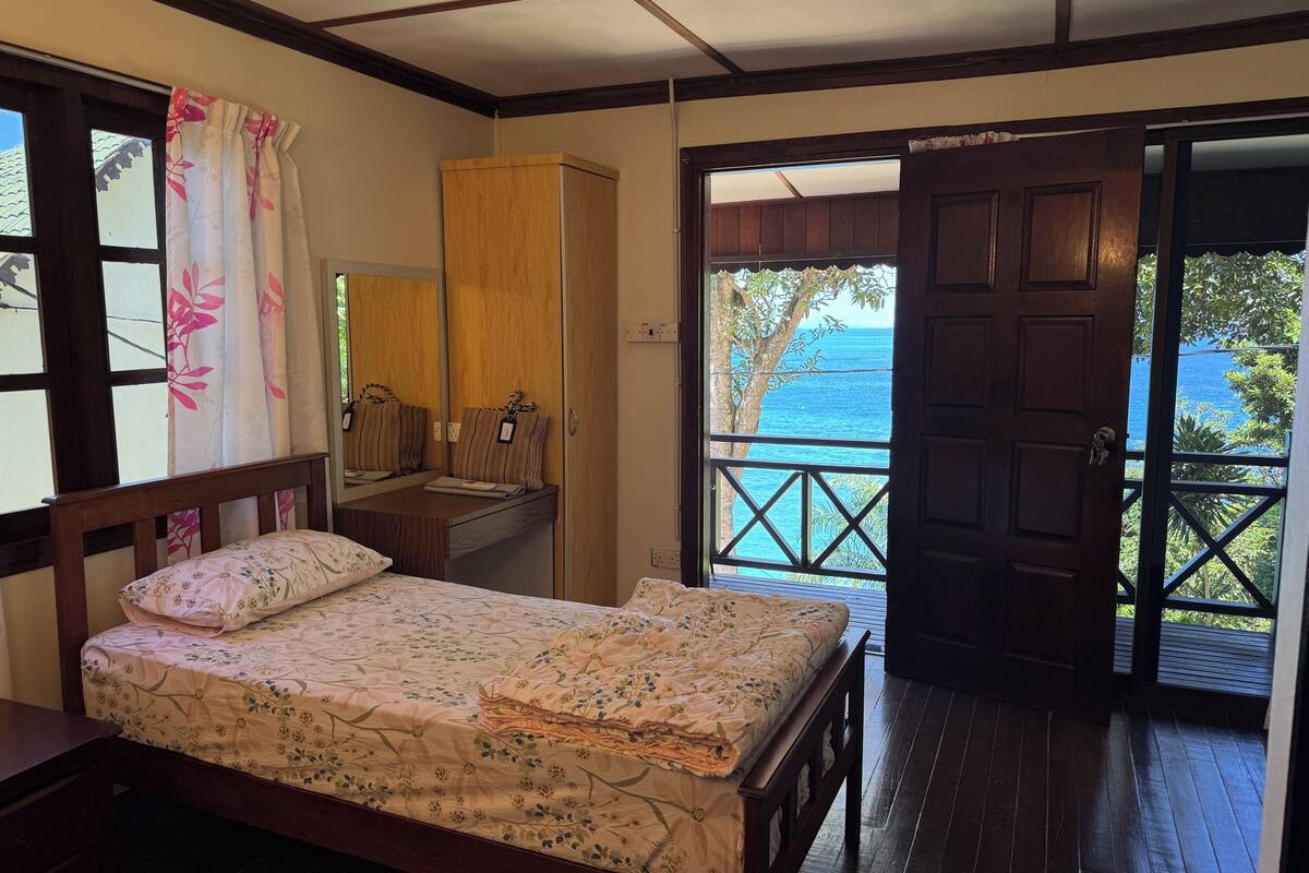 A bedroom with a bed, a wooden wardrobe, a desk with a mirror, and a view of the sea through an open door.