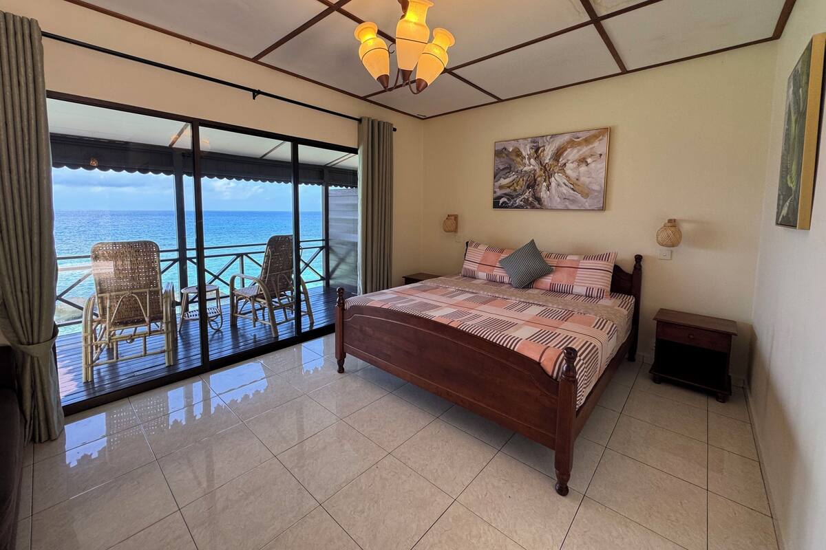 A bedroom with a bed, a nightstand, a painting, a ceiling light, and a view of the ocean through large windows.
