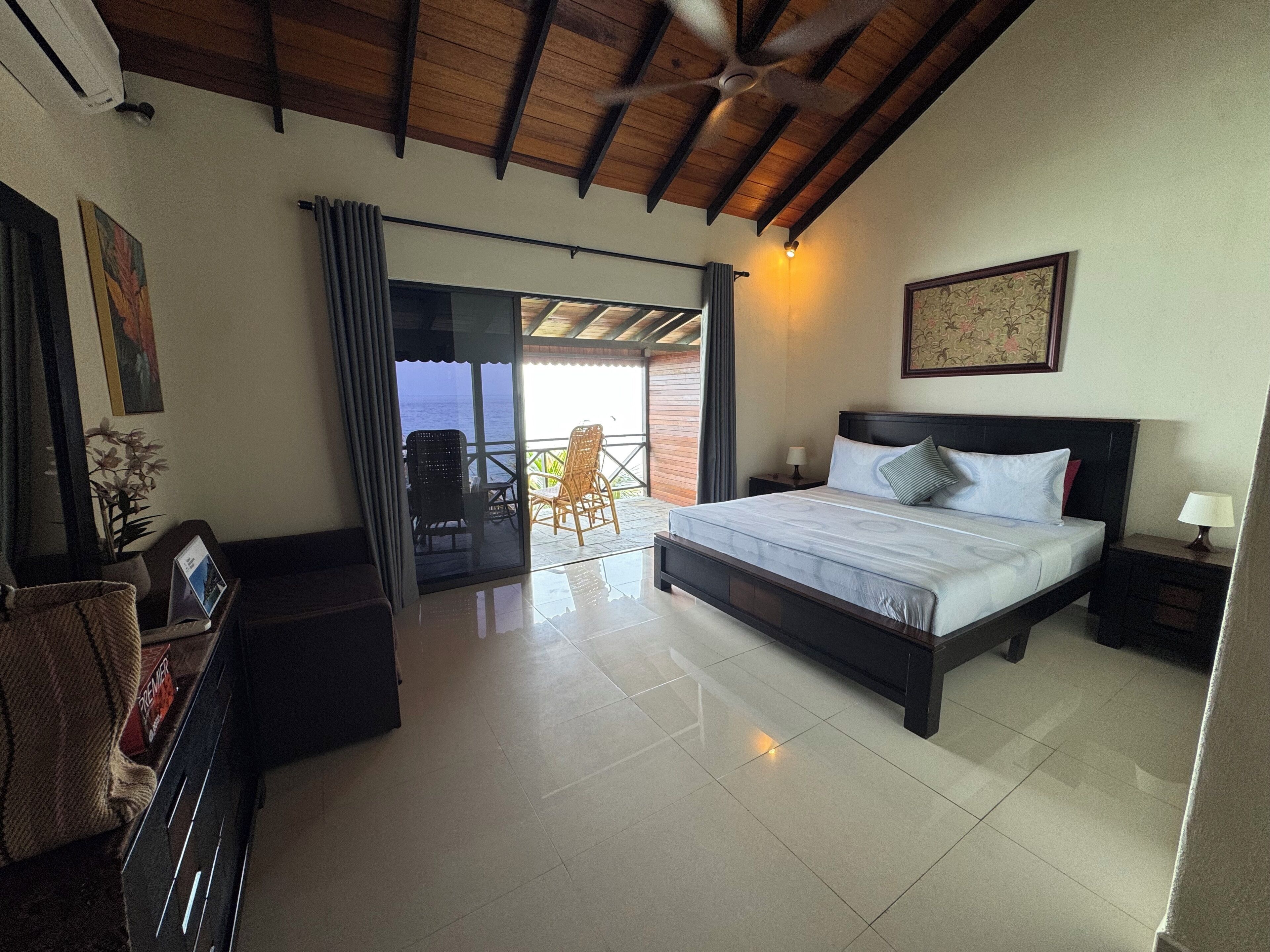 Una camera da letto con un letto, un comodino, una scrivania, una sedia, una finestra con tende e vista sul mare.