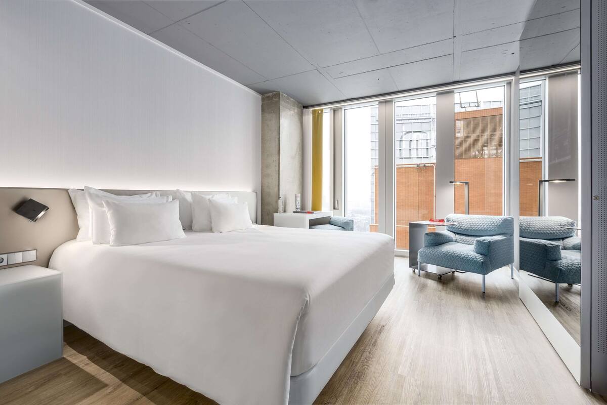 A modern hotel room with a large bed, a nightstand, a seating area with chairs, and a view through a glass door.