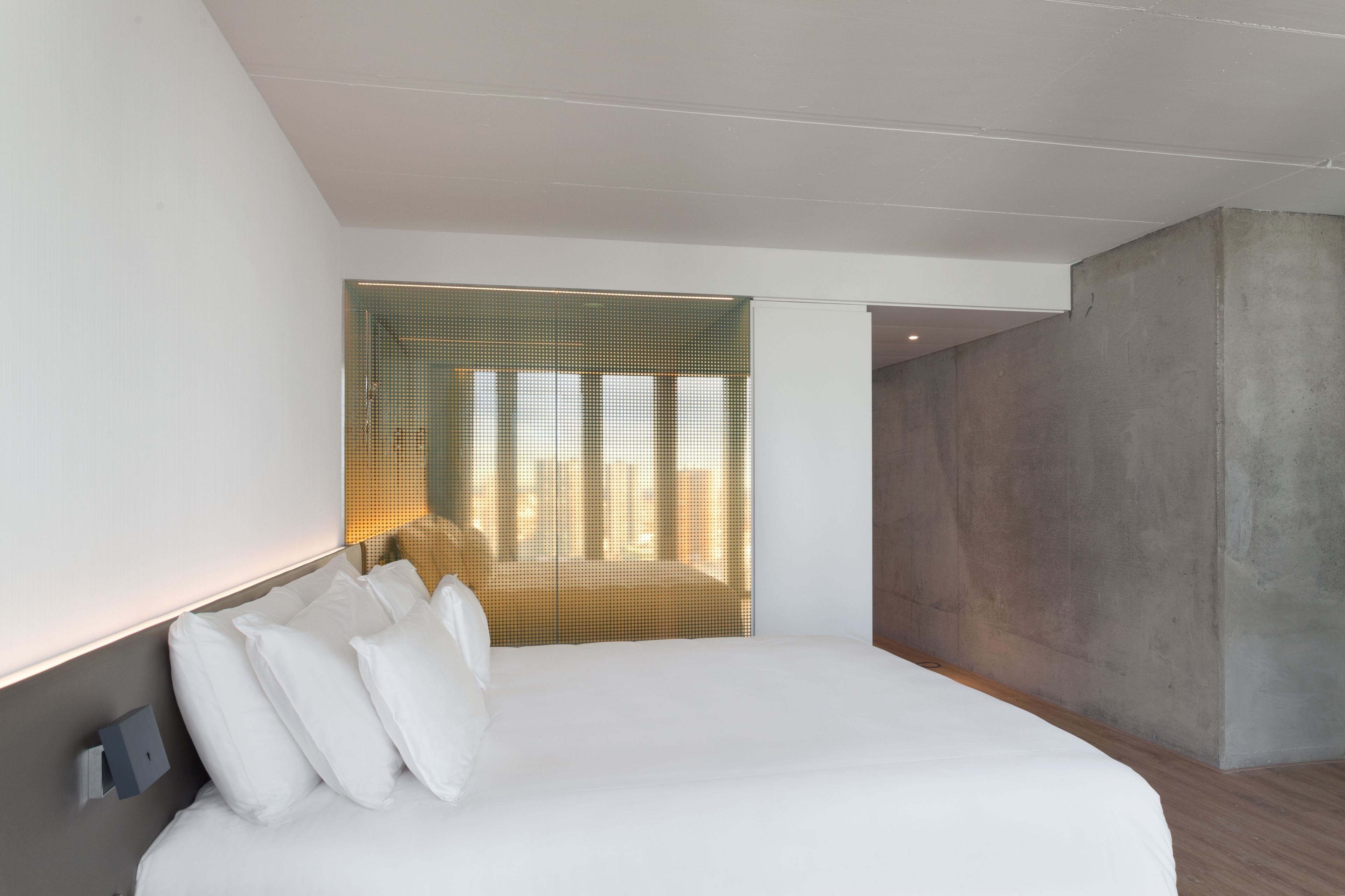A modern hotel room with a large bed, a concrete accent wall, and a unique textured window blind.