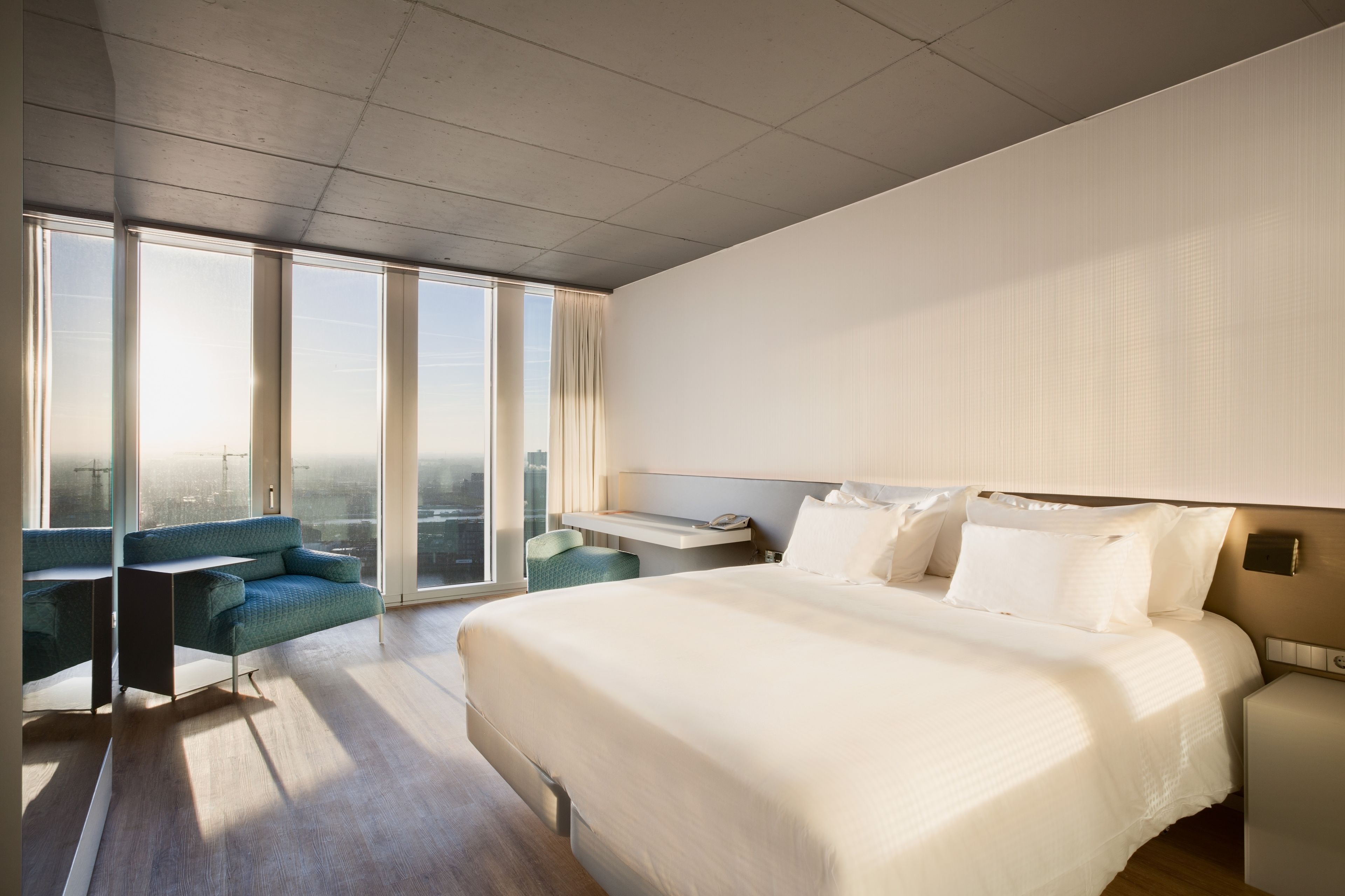 A modern hotel room with a large bed, a desk, a chair, and a view of the city through large windows.