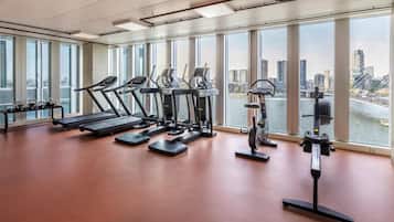 A gym with treadmills, ellipticals, and a rowing machine, large windows overlooking a cityscape and a river.