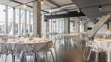 A spacious dining area with round tables and white chairs, large windows offering views of the city, and exposed ductwork on the ceiling.