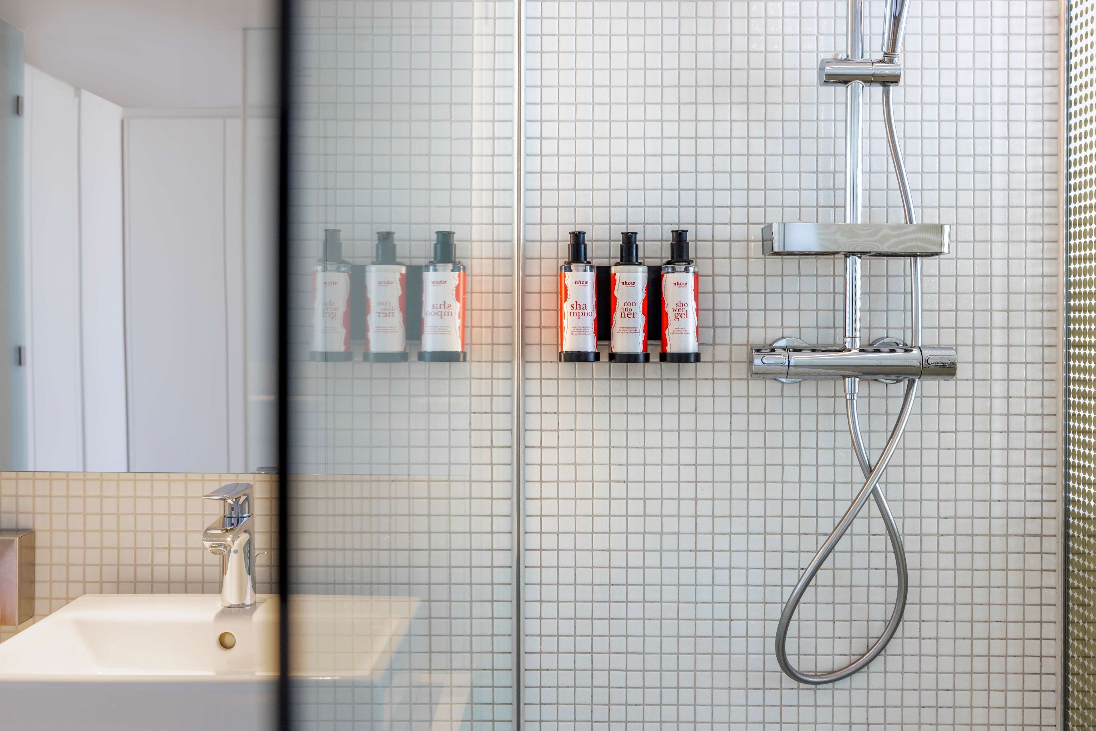 Un baño con un lavamanos blanco, un espejo y una ducha con cabezal de ducha manual. Hay tres botellas de champú y acondicionador en la pared.