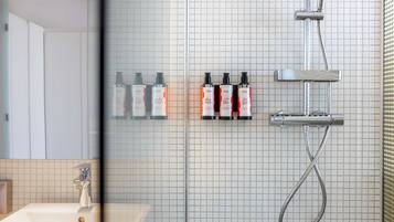 Un baño con un lavamanos blanco, un espejo y una ducha con cabezal de ducha manual. Hay tres botellas de champú y acondicionador en la pared.