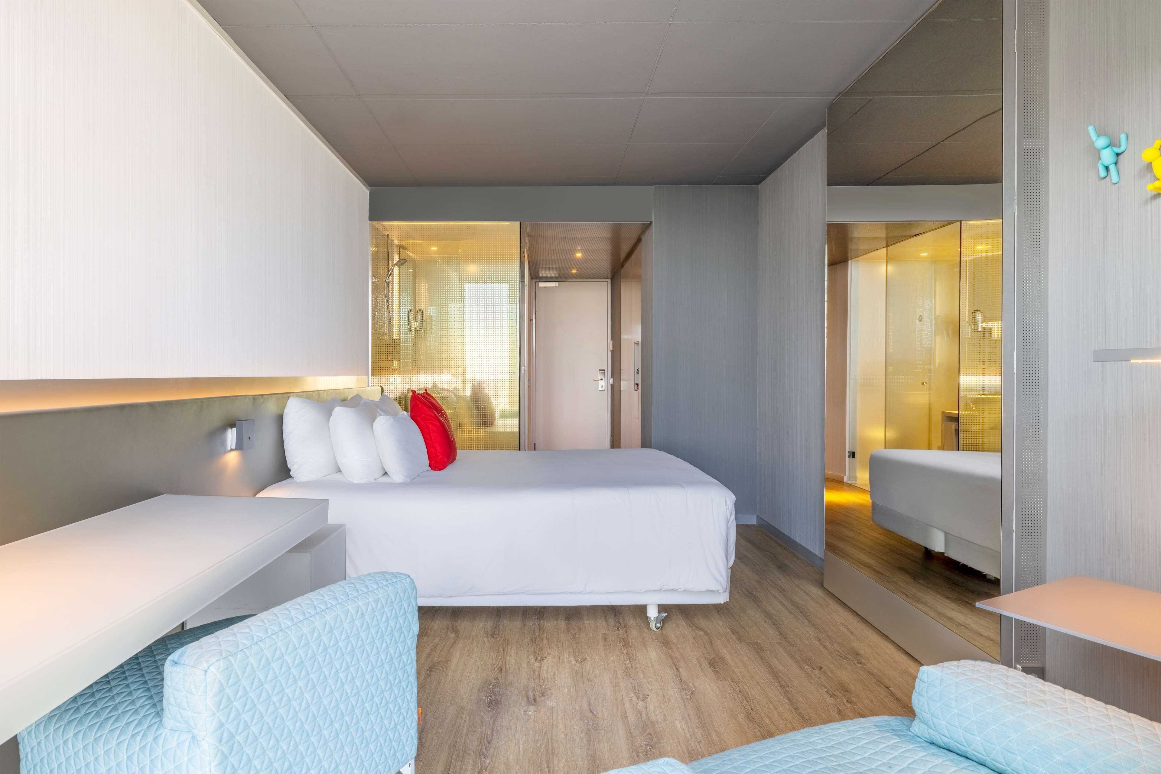 A modern hotel room with a large bed, a bedside table, a chair, and a glass-enclosed bathroom.