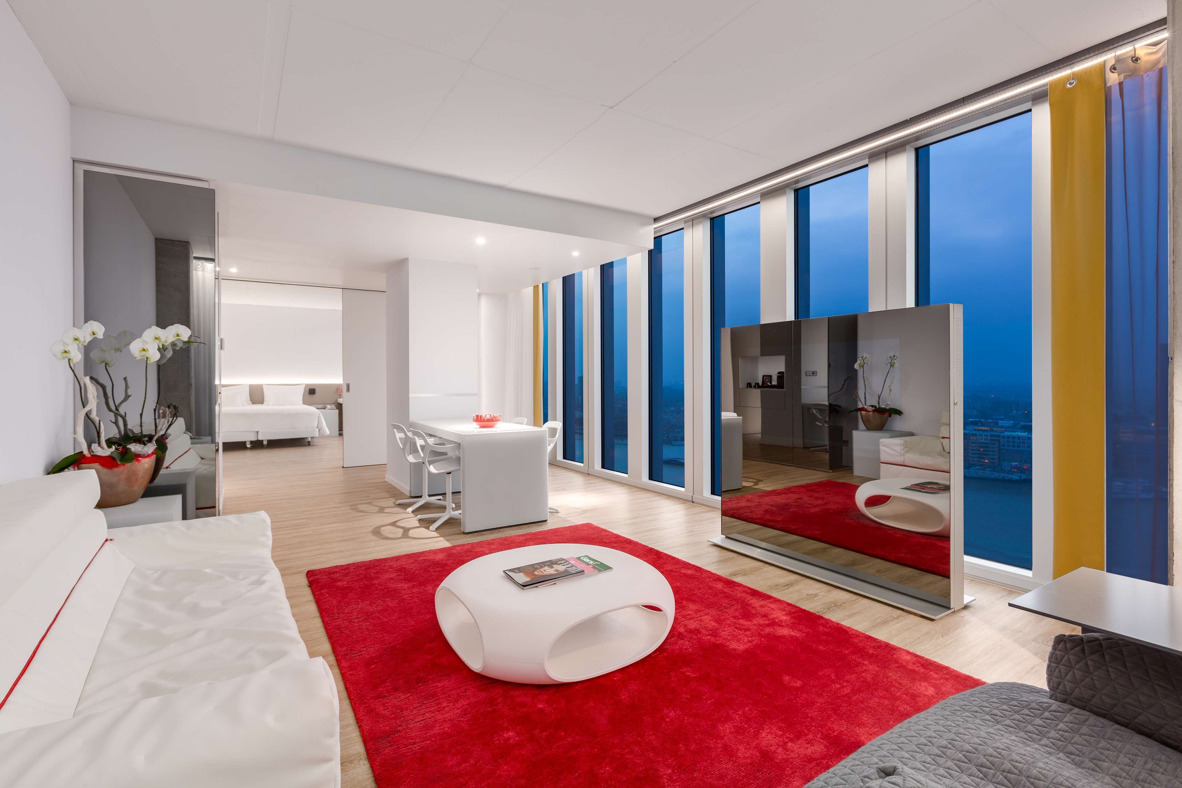 Een moderne hotelkamer met een groot raam, een rood vloerkleed, een witte bank, een witte fauteuil, een klein wit tafeltje en een spiegel.