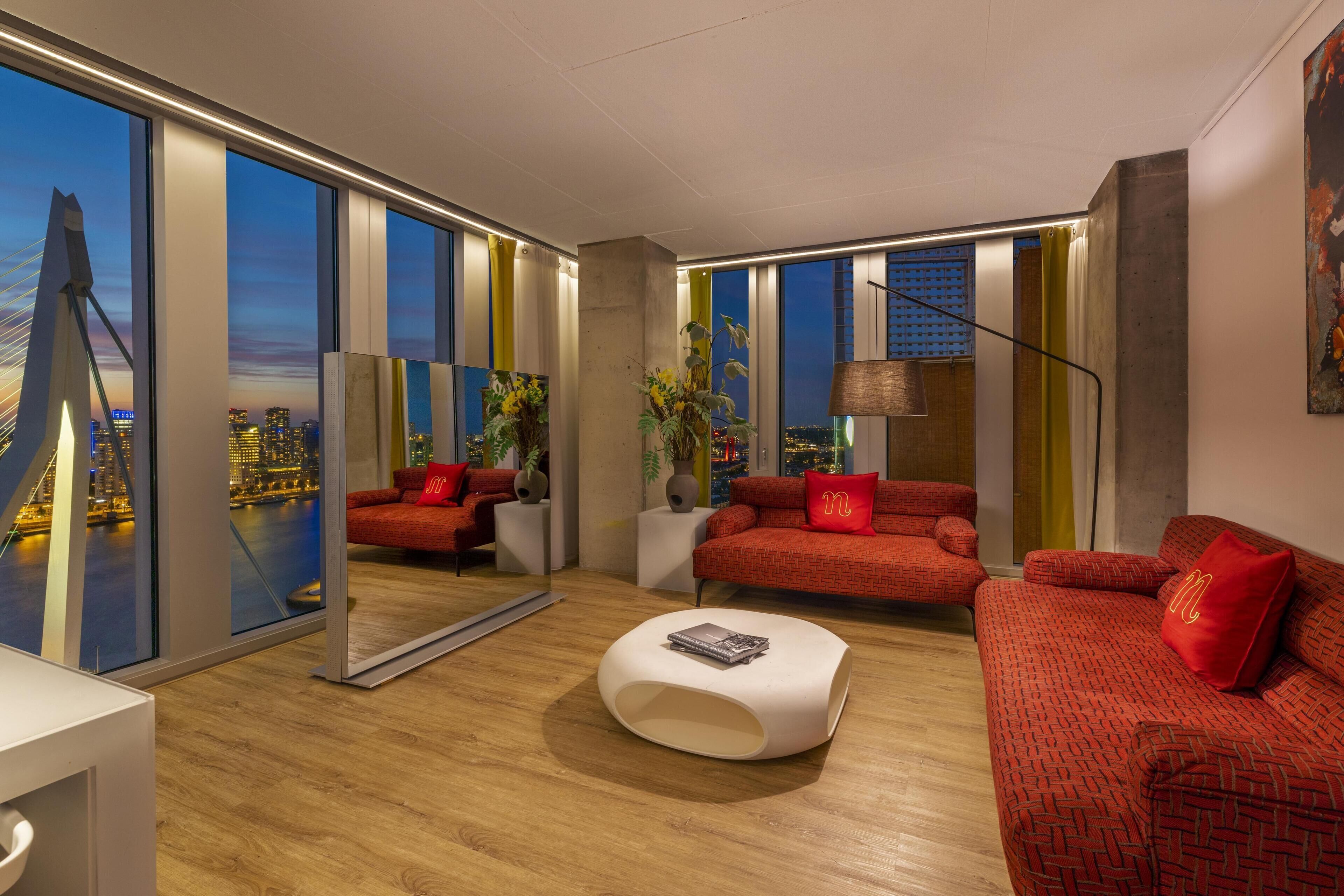 Una sala de estar moderna con ventanas amplias, sofás rojos, una mesa de centro blanca y vistas a la ciudad.