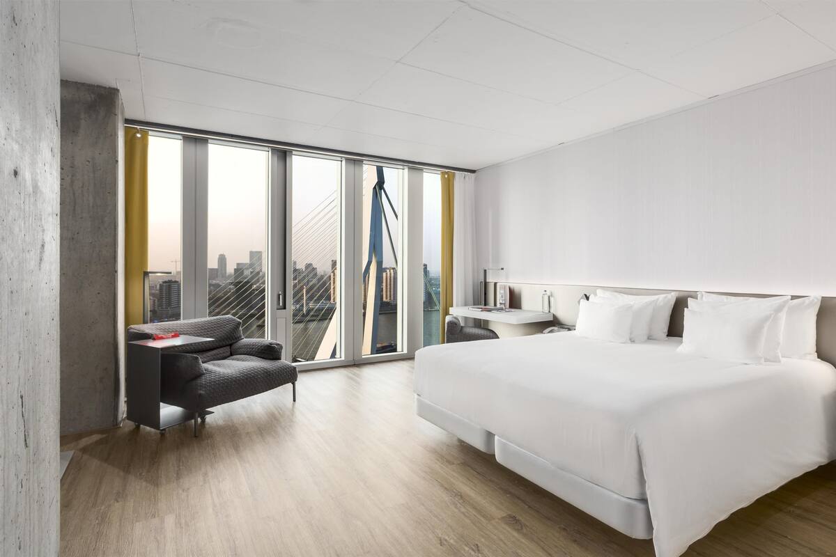 A modern hotel room with a large bed, a desk, a sofa, and a view of the city through floor-to-ceiling windows.