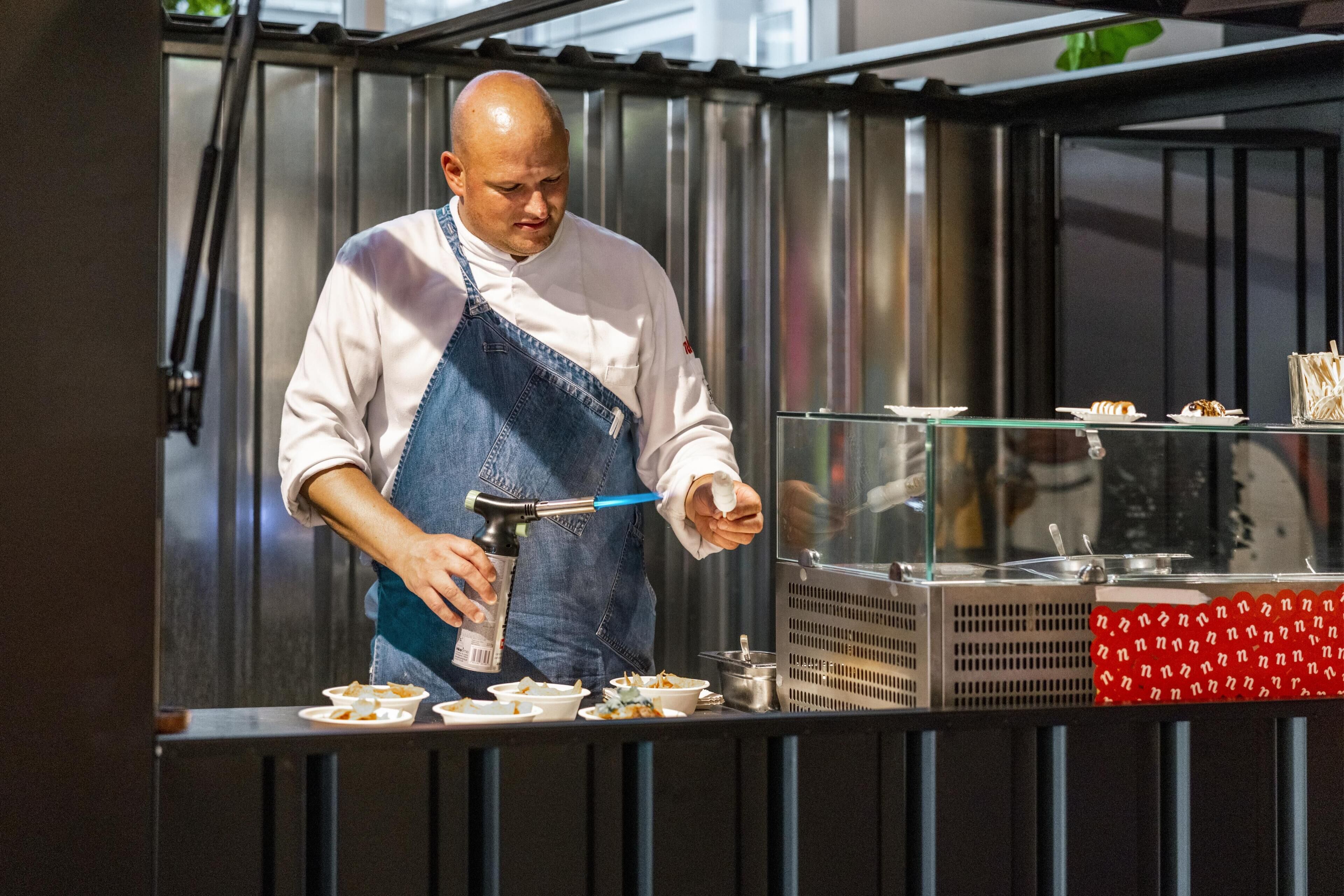 Een kok in een witte blouse en een spijkerimbi zorgend voor eten aan een toonbank met meerdere kommen en een grill.