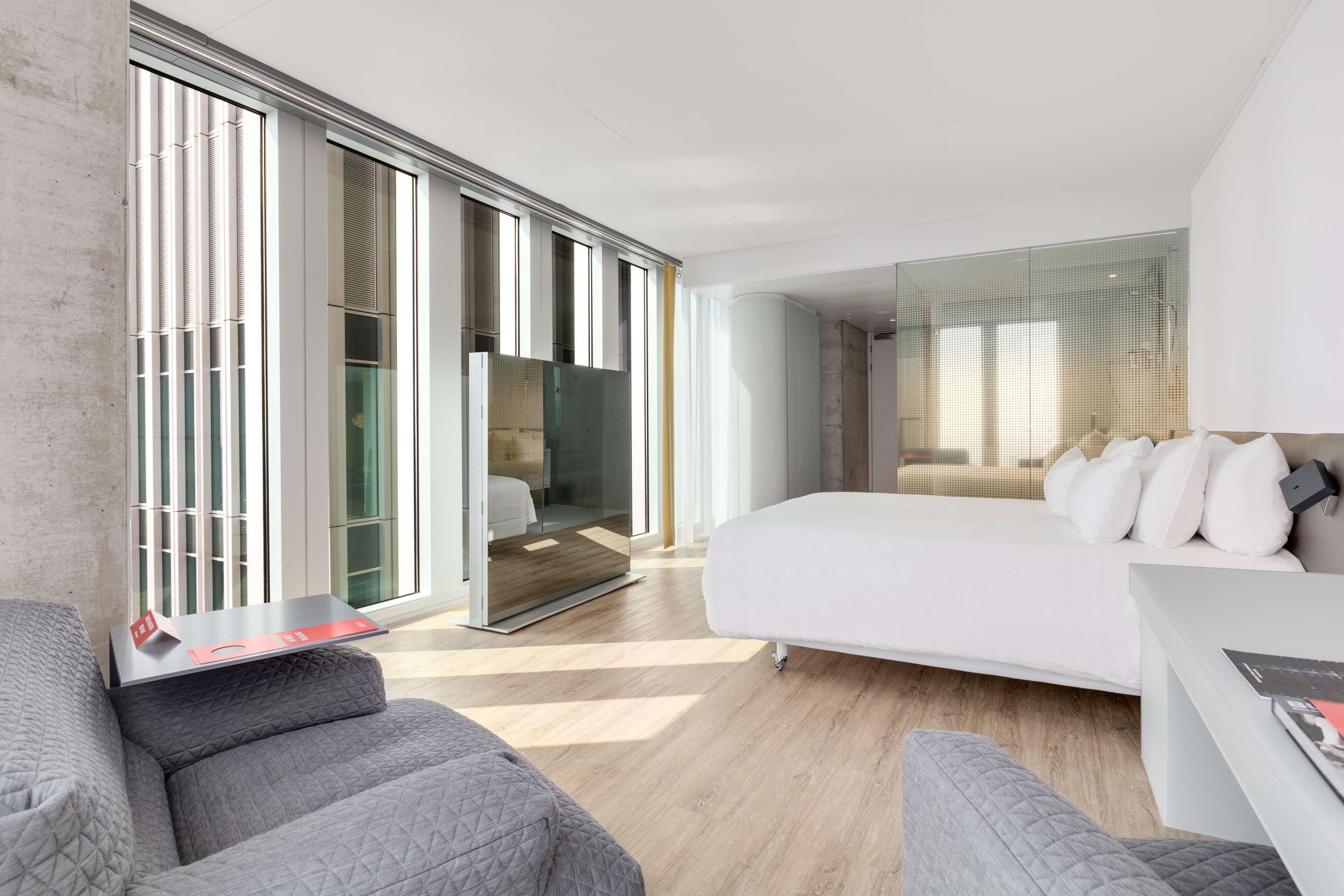 A modern hotel room with a large bed, a seating area with a sofa and armchair, a small table, and a glass partition separating the room from the hallway.