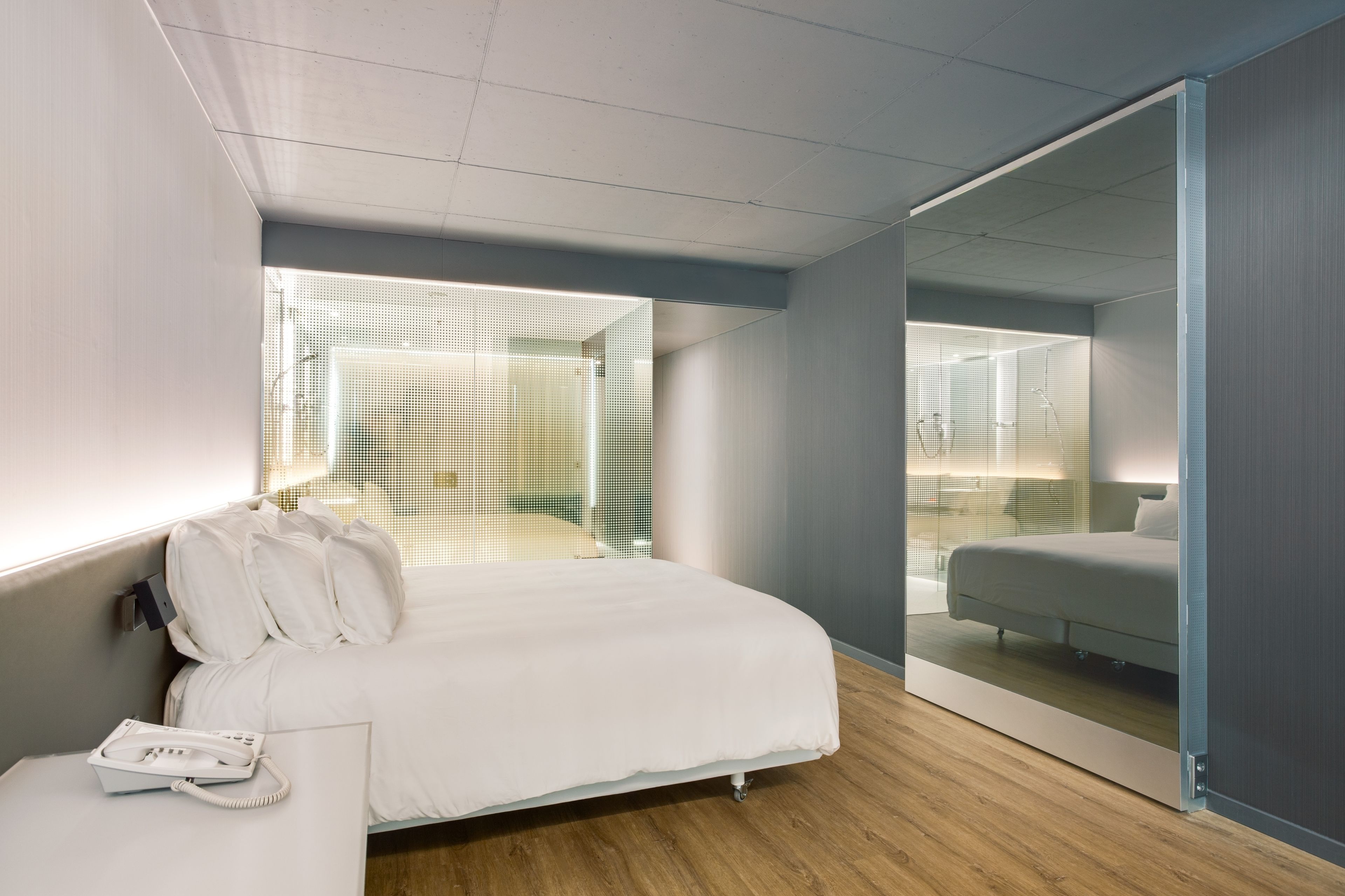 A modern hotel room with a large bed, a bedside telephone, and a view through a glass partition to a bathroom.