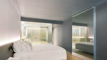 A modern hotel room with a large bed, a bedside telephone, and a view through a glass partition to a bathroom.