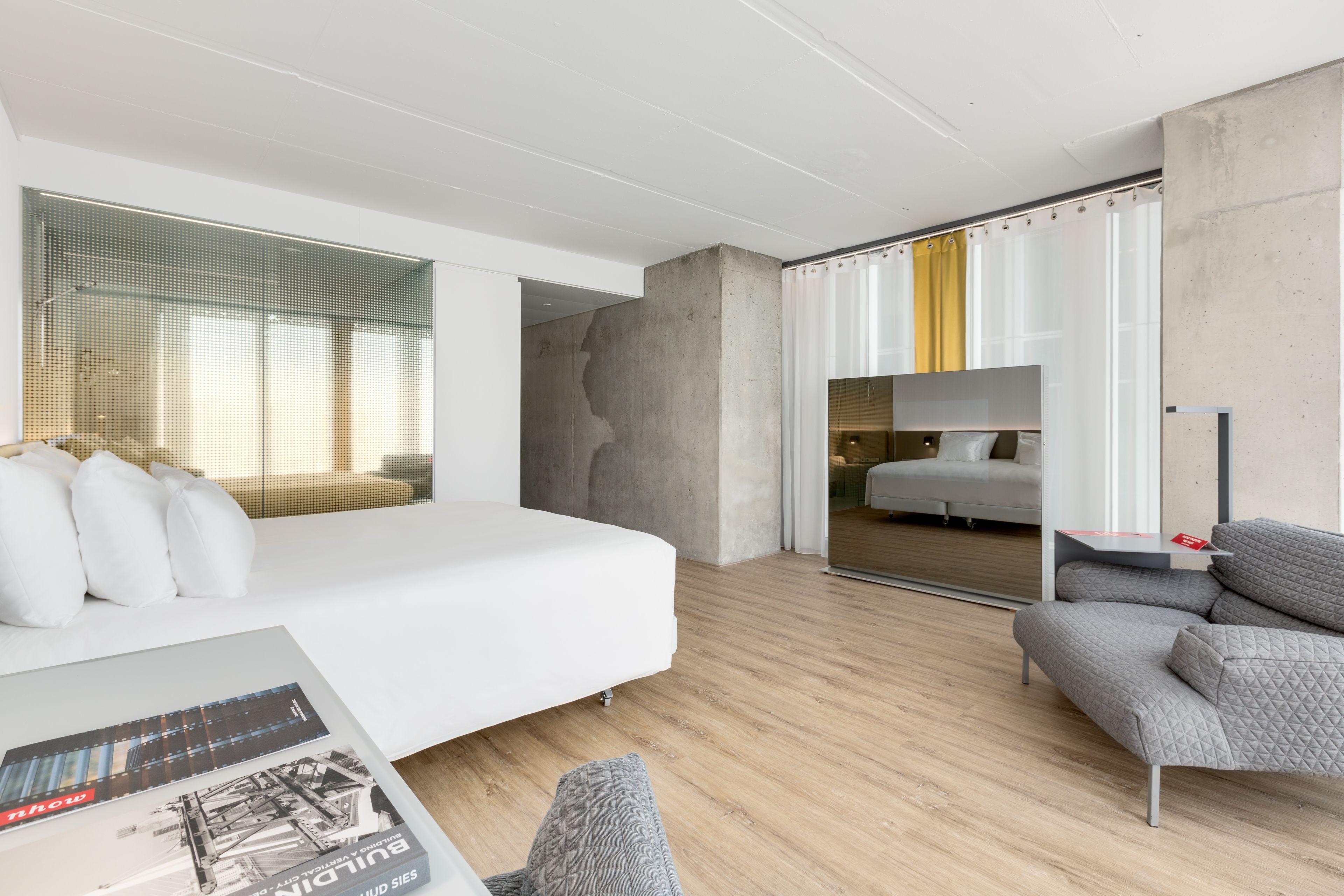 A modern hotel room with a large bed, a grey sofa, and a large window with curtains.