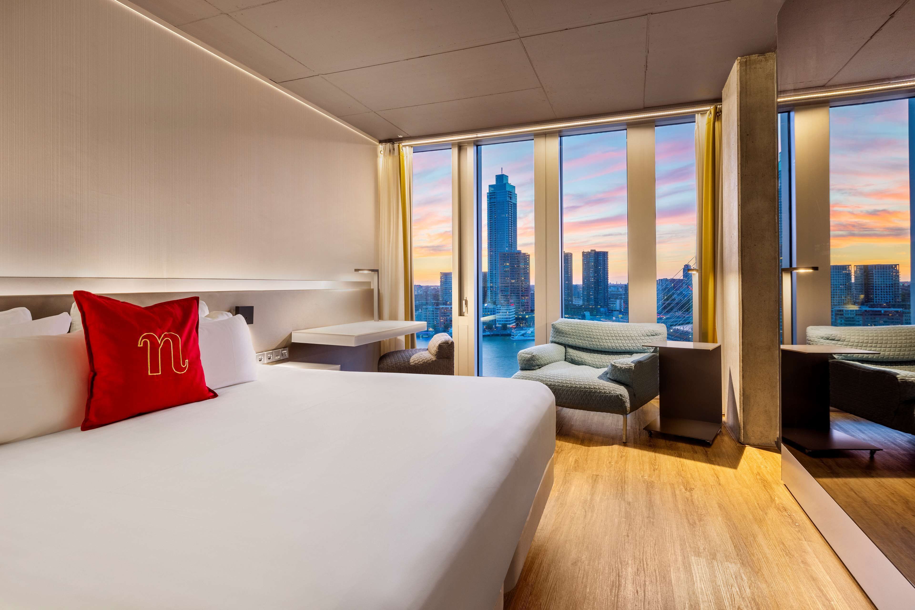 A hotel room with a large bed, a desk, and a view of the city skyline at sunset.