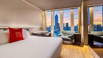 A hotel room with a large bed, a desk, and a view of the city skyline at sunset.