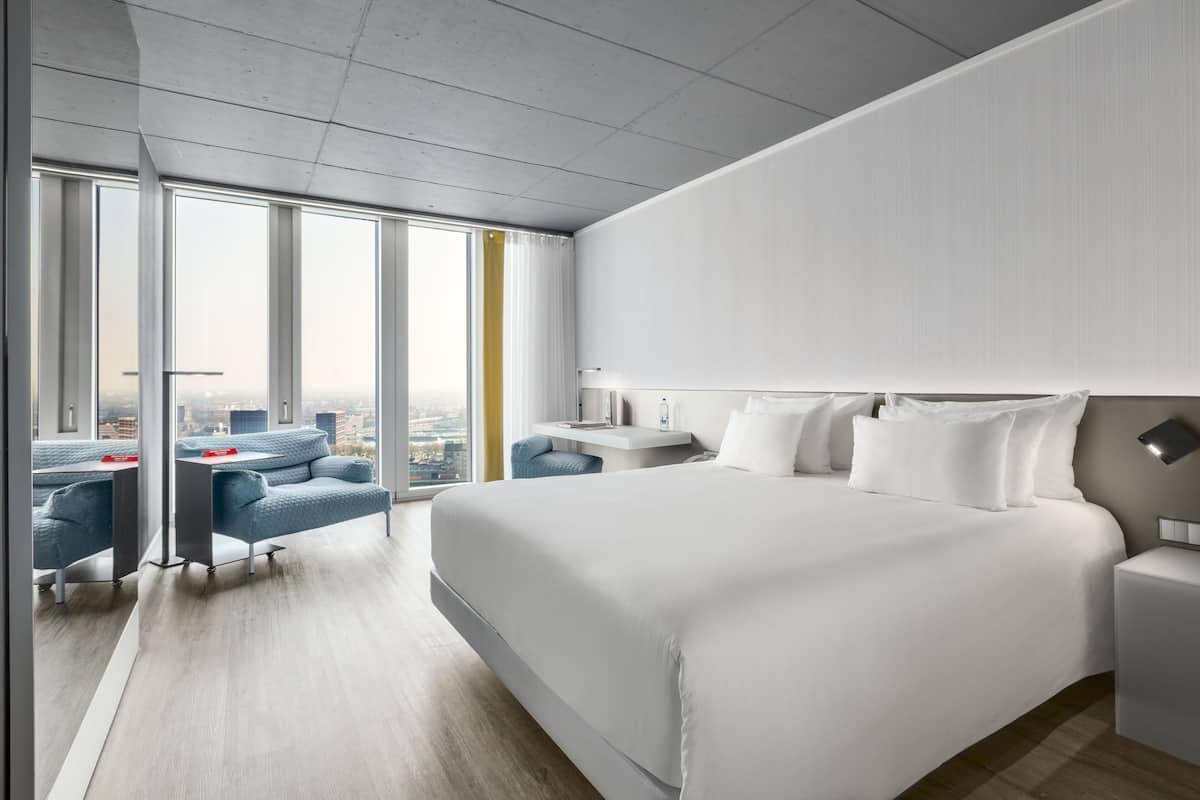 A modern hotel room with a large bed, a desk, and a chair. There are large windows offering a city view.
