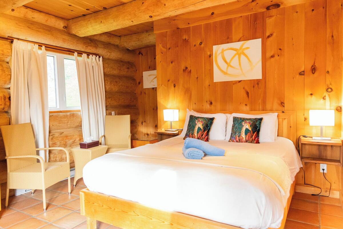 Ein Zimmer in einer Holzhütte mit einem Bett, zwei Nachttischen, einem Sessel und einem Fenster mit Vorhängen.
