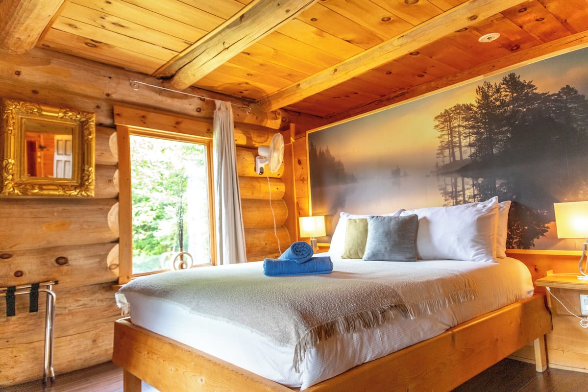 Ein Schlafzimmer einer Holzhütte mit einem Bett, einem Nachttisch, einem Wandventilator, einem Fenster mit Vorhängen und einem gerahmten Bild.