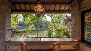 Una mesa de madera con dos sillas, una lámpara colgante y vistas a un jardín con árboles y un estanque.