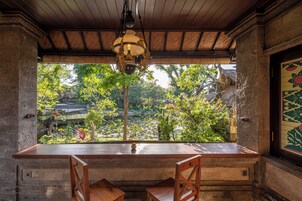 Mesa de madeira com duas cadeiras, luminária pendente e vista para um jardim com árvores e lago.
