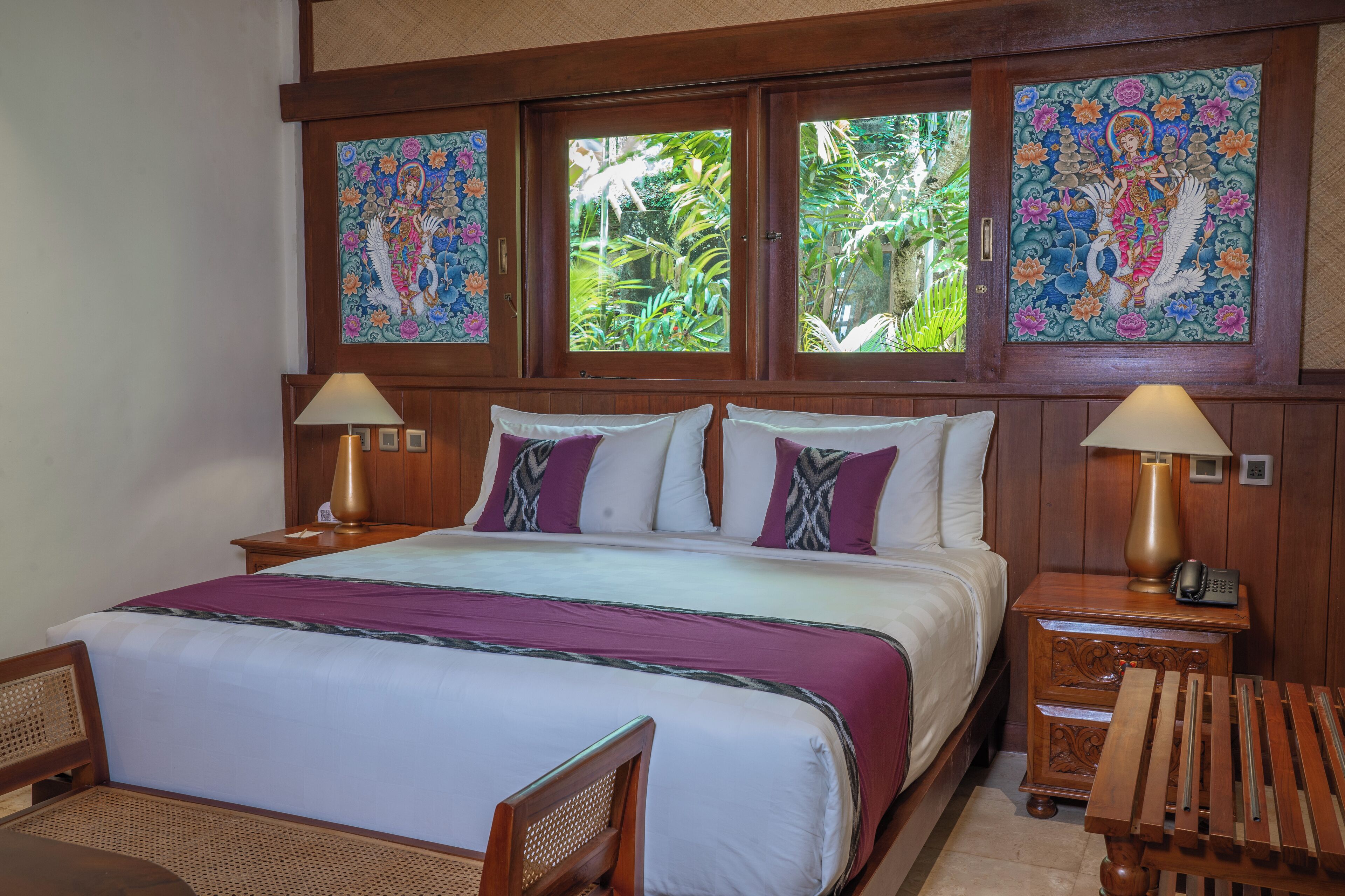 A hotel room with a large bed, two bedside tables with lamps, a wooden chair, and a view of greenery through the windows.