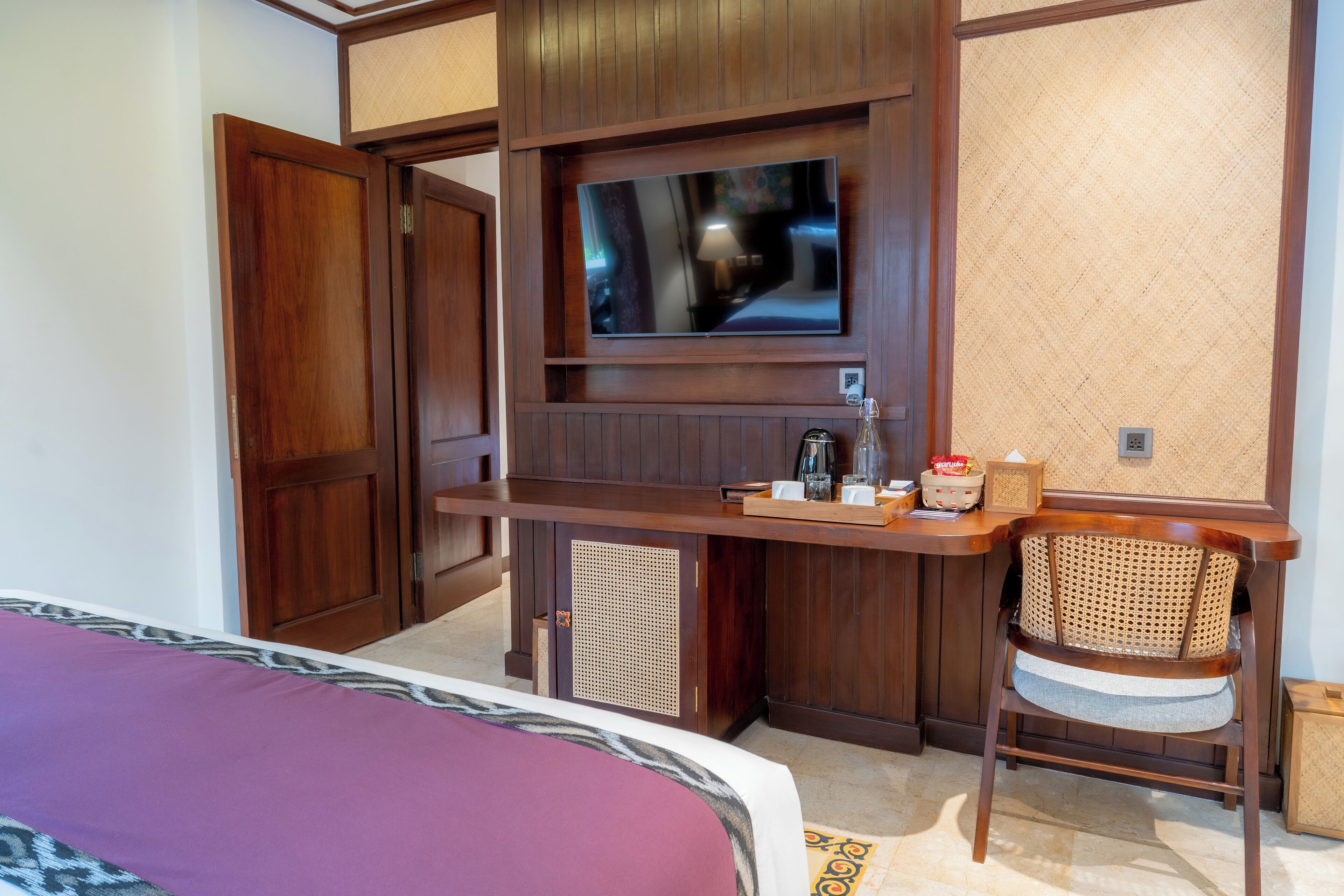 Une chambre d’hôtel avec un bureau en bois, un téléviseur fixé au mur, un fauteuil en osier et un lit recouvert d’une couverture violette.