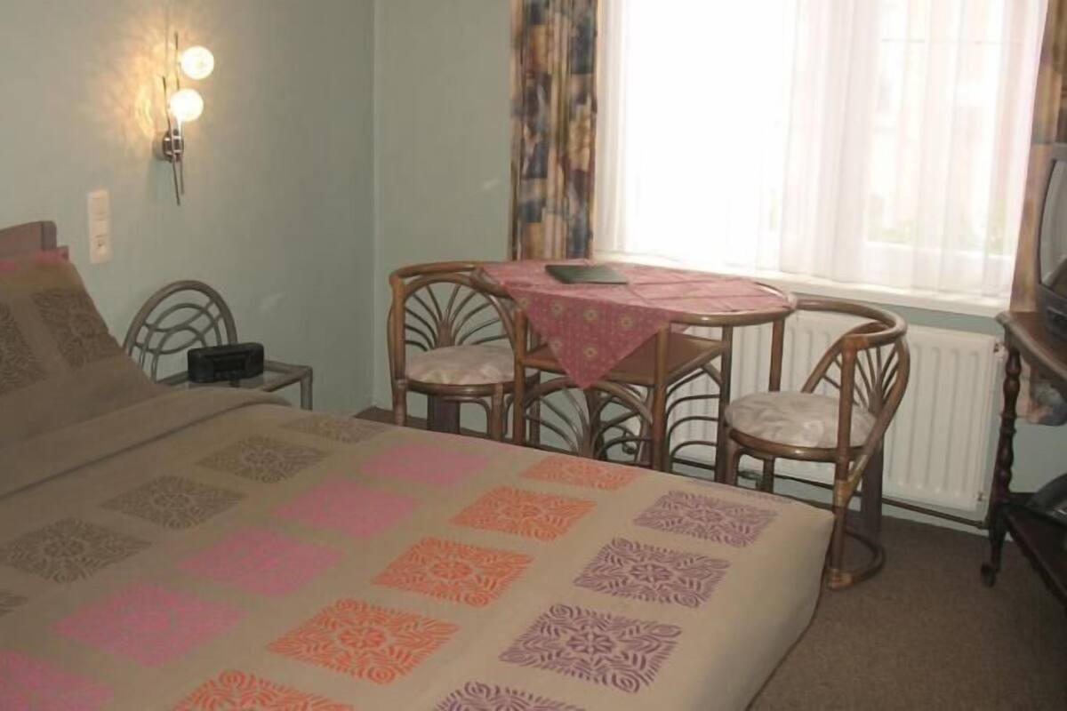 A bedroom with a bed, a small table with chairs, a wall-mounted light, and a window with curtains.