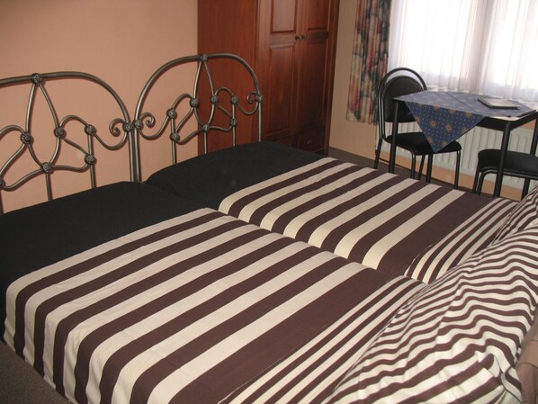 Una cama doble con ropa de cama a rayas, un cabecero de metal, una puerta de madera, una mesita con mantel y una silla.