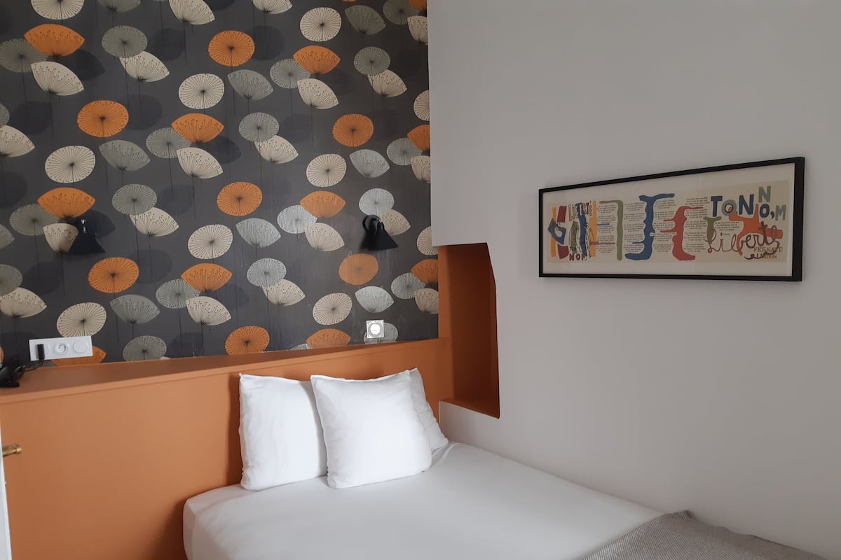 Hotellihuone, jossa on sänky, valkoiset tyynyt, sängynpääty, kuvioidulla tapetilla päällystetty seinä ja kehystetty taideteos.