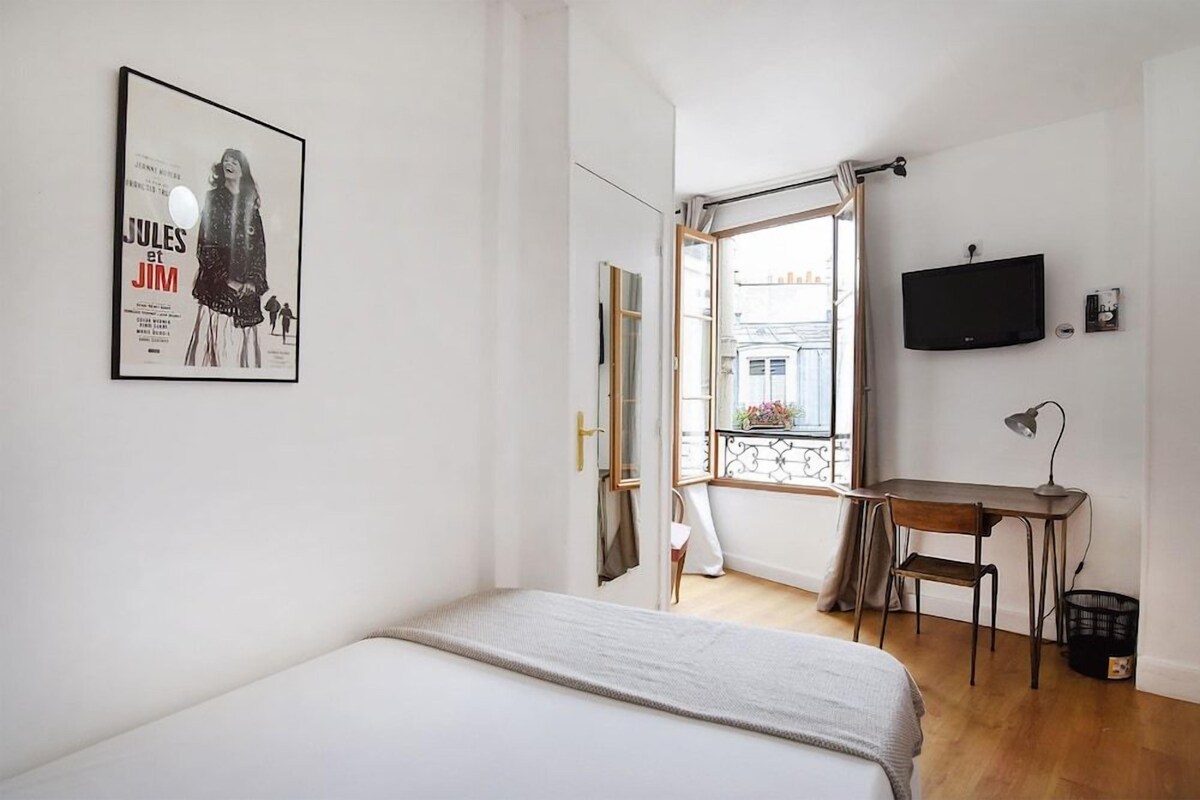 A bedroom with a bed, a desk, a chair, a TV, and a window with a view of buildings.