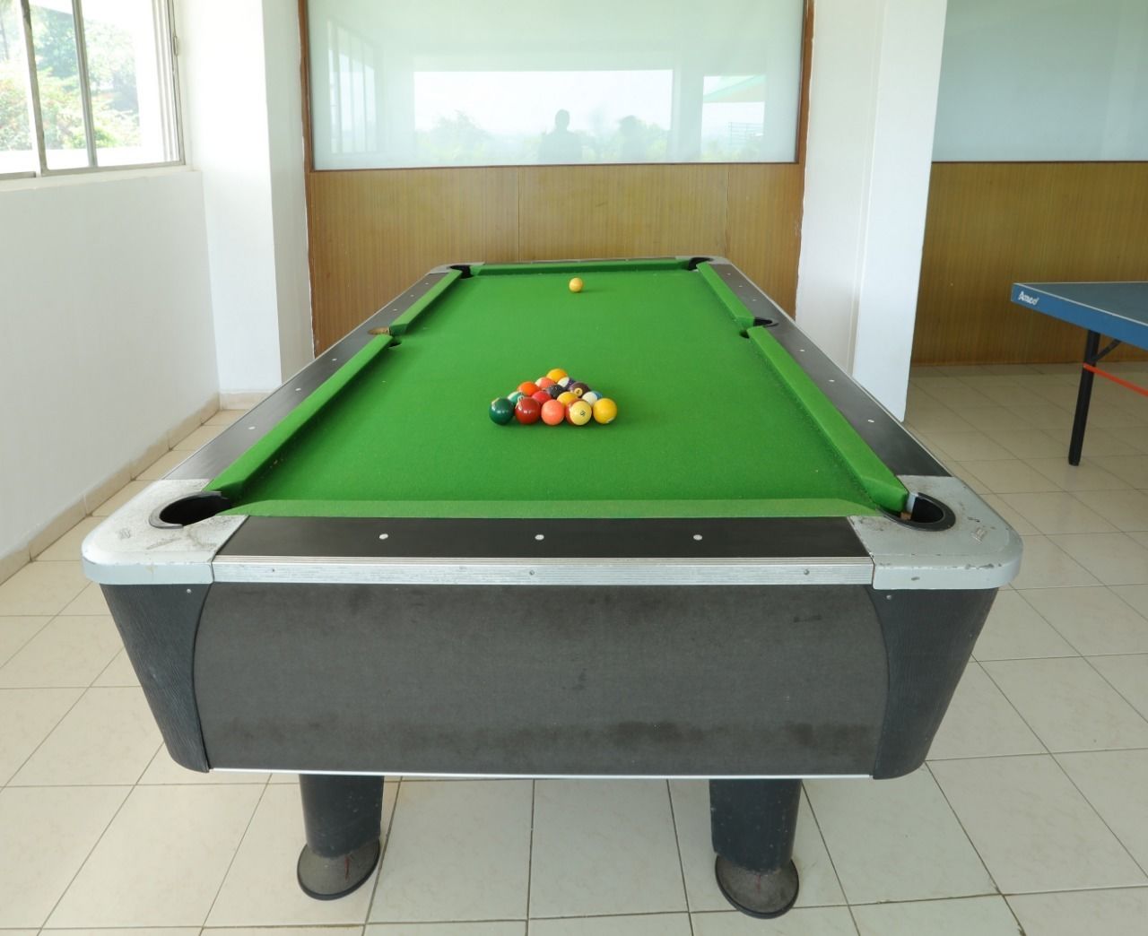 Una mesa de billar con bolas en una habitación con suelo de baldosas y un ventanal.
