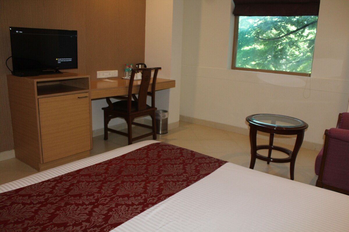 Ein Hotelzimmer mit einem Bett, einem Schreibtisch mit Computer, einem Stuhl, einem kleinen Tisch und einem Fenster mit Blick auf Bäume.