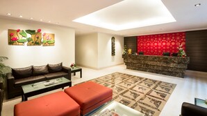 Ein modernes Wohnzimmer mit einer Couch, Hocker und Couchtisch. Im Hintergrund ist eine Rezeption mit einer roten Wand mit Muster zu sehen.