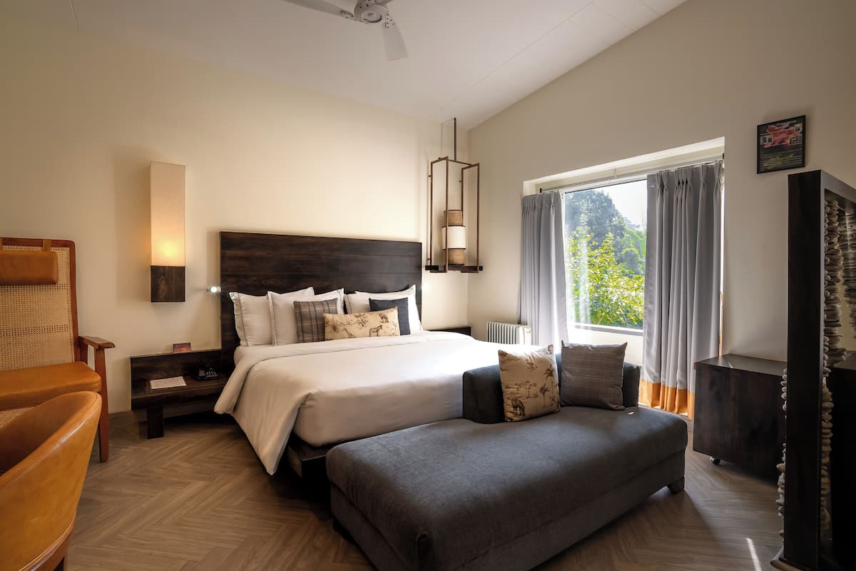 A hotel room with a large bed, a sofa, a wooden nightstand, a lamp, a ceiling fan, and a window with curtains.