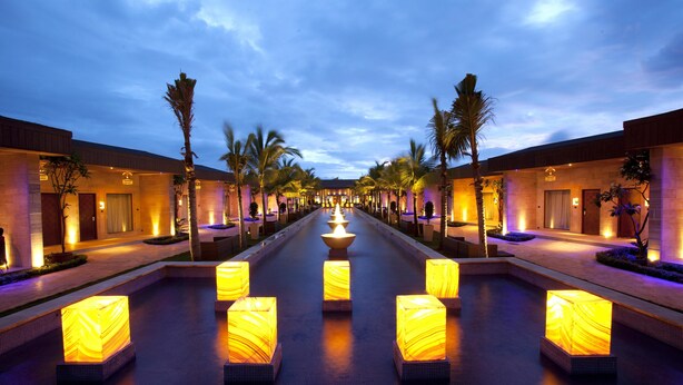 A hotel corridor with a central water feature, palm trees, and illuminated square lights. - Della Resorts (Mawal)