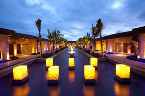 A hotel corridor with a central water feature, palm trees, and illuminated square lights. - Della Resorts (Mawal)
