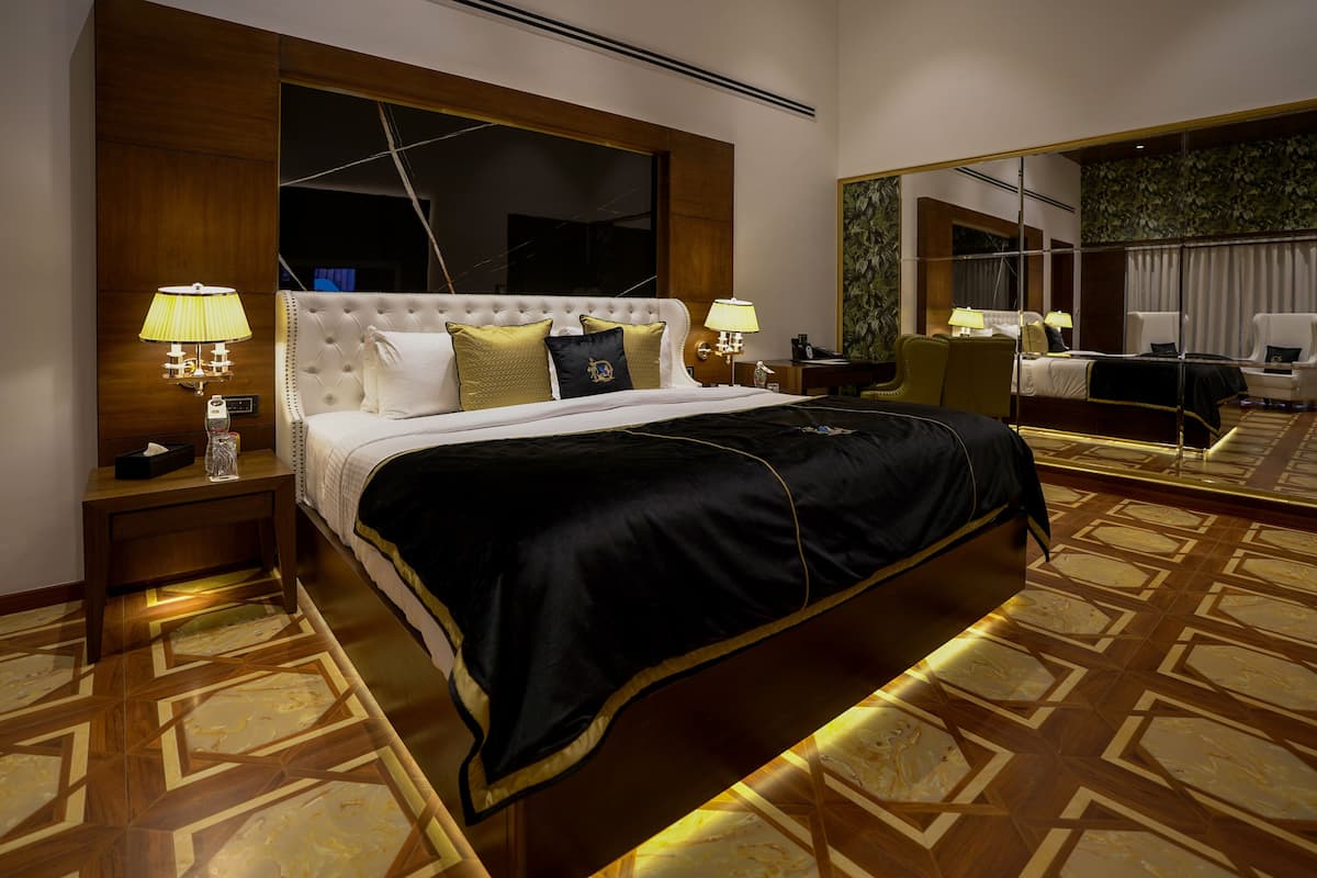Quarto de hotel com uma cama grande, duas mesas de cabeceira, um criado-mudo, um espelho e um tapete estampado.