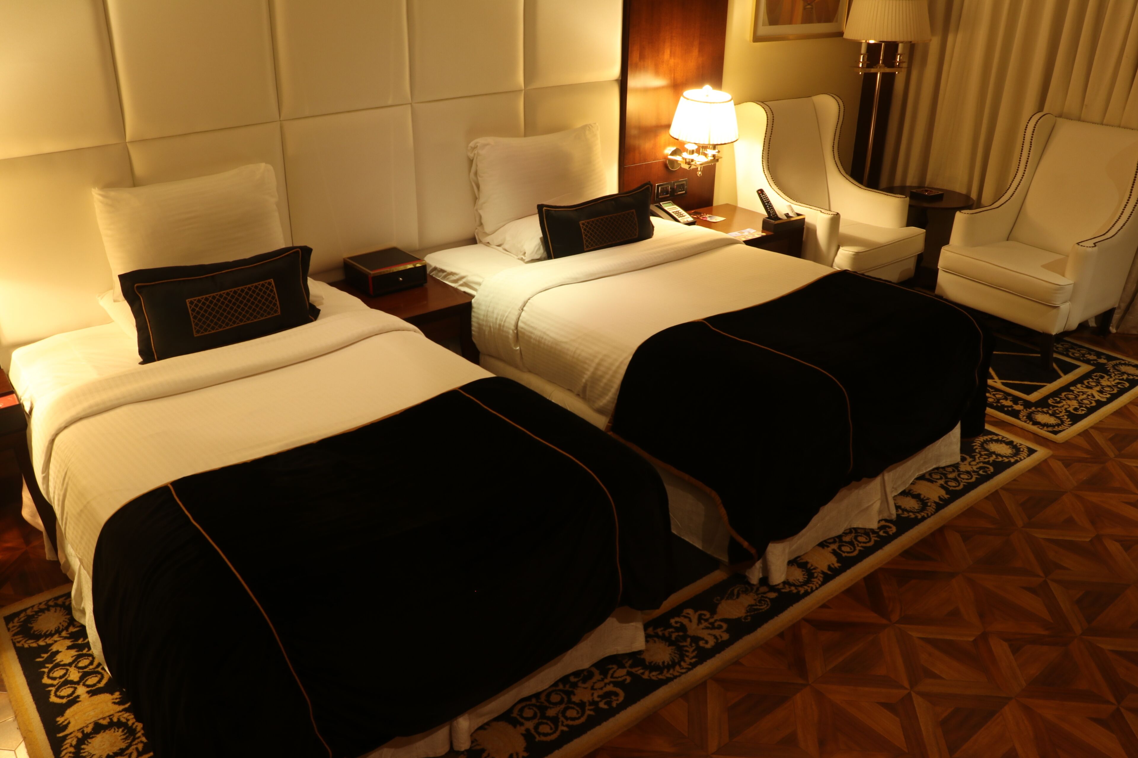 A hotel room with two beds, a nightstand with a lamp, a chair, and a patterned rug.