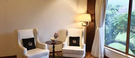 Una habitación con dos sillones blancos, una mesita redonda con una lámpara y vistas al verdor a través de una ventana.