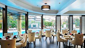 A modern dining area with round tables and chairs, large windows with a view of a pool and greenery. - Della Resorts (Mawal)
