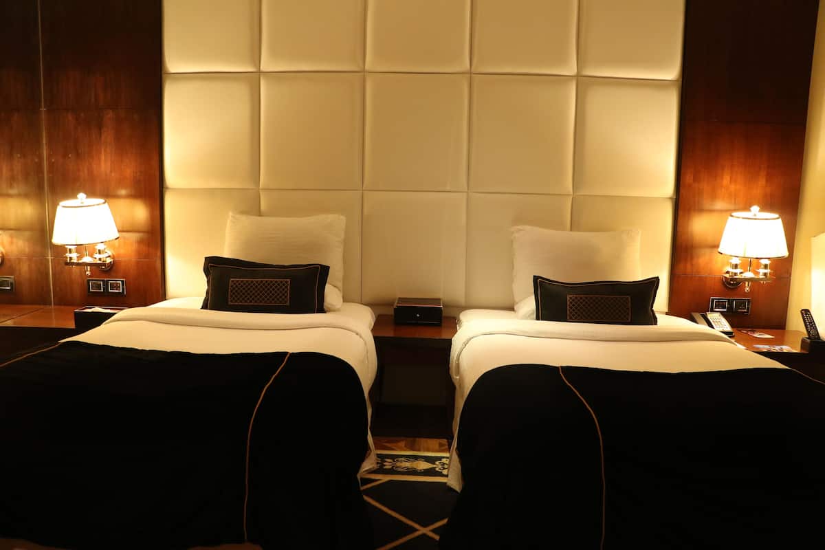 Quarto de hotel com duas camas, cada uma com travesseiro e mesa de cabeceira com abajur.