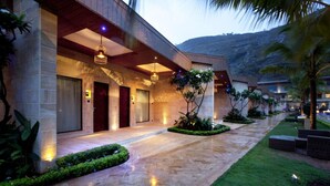 A modern hotel entrance with a stone facade, a covered walkway, and well-maintained landscaping. - Della Resorts (Mawal)