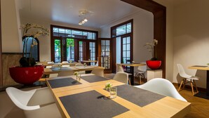 Un comedor moderno con mesas de madera, sillas blancas, ventanas amplias y plantas en macetas.