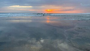 Una playa con arena mojada que refleja el cielo, una persona caminando y el océano al fondo.