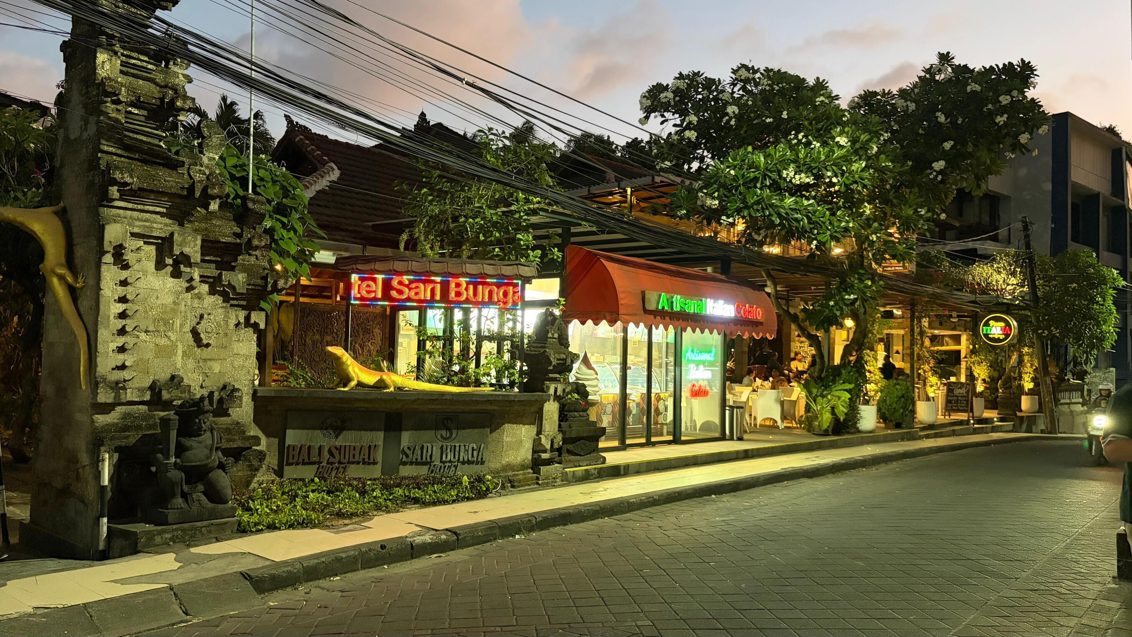 A street with a restaurant named 'Tel Sari Bung' and another called 'Atsanal Italian Gelato.' There is a statue of a crocodile and a sign for 'Bali Subur Hotel.'