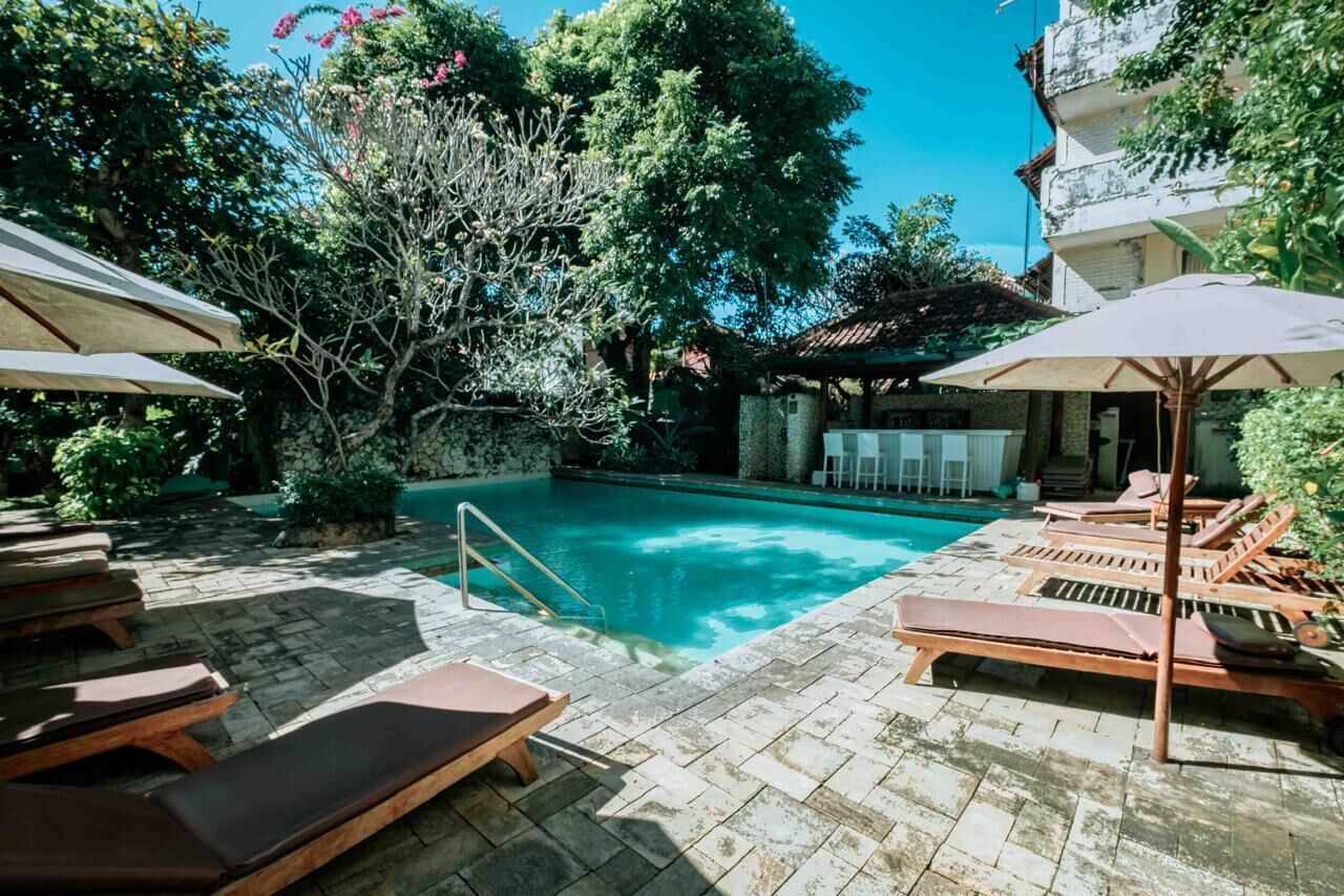 Piscina con sillones de descanso y sombrillas, rodeada de vegetación y un edificio.