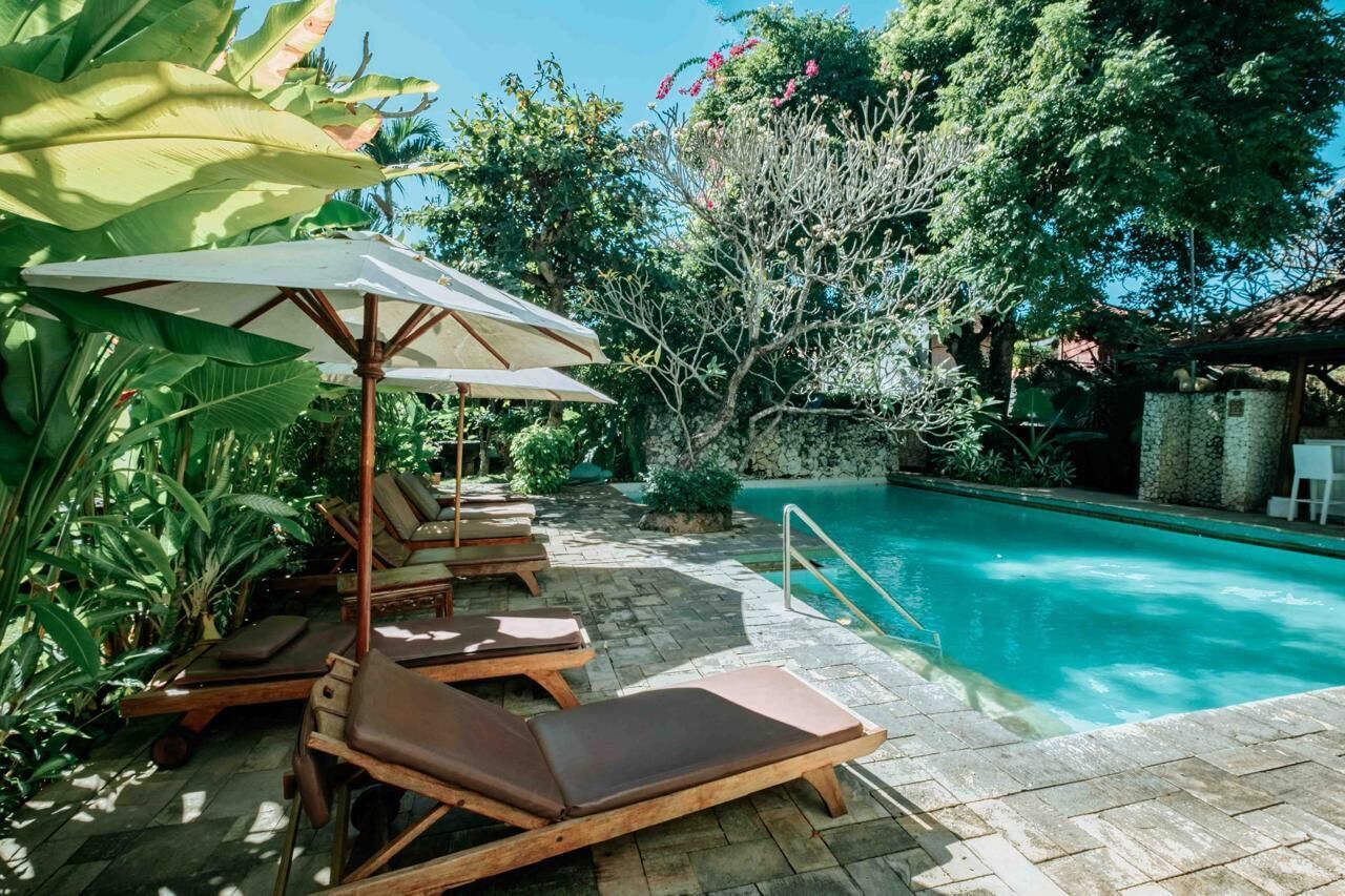 A poolside area with lounge chairs and an umbrella, surrounded by lush greenery and a clear blue pool.